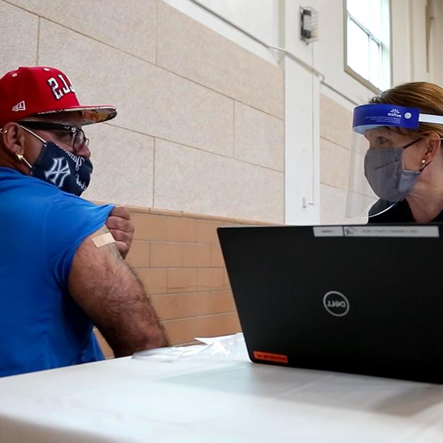 凯萨琳页面, 戴着面具和盾牌, 是在笔记本电脑旁边，一个男人正在撩起袖子，展示他接种疫苗的地方的绷带. 