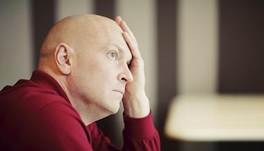 A bald man in a red shirt holds his hand to his forehead, looking preoccupied.